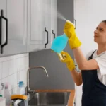 Woman in white-blue uniform and yellow gloves cleaning kitchen cabinet holding a blue bottle in her hand.