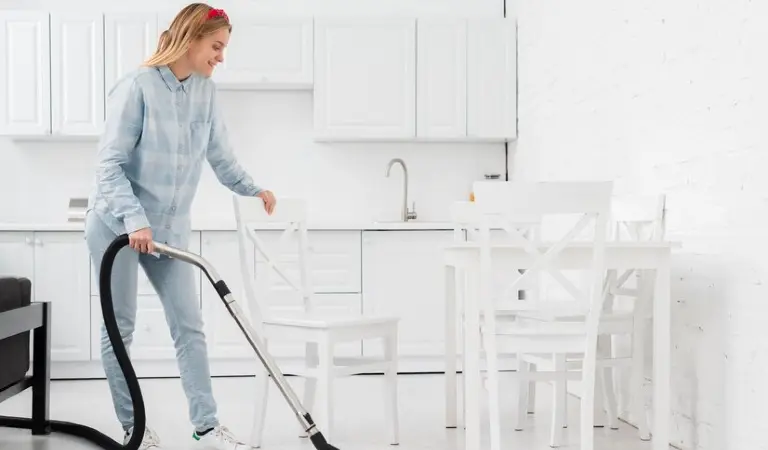 A lady in check shirt holding a vacuum cleaner in her hand with white chairs around her.