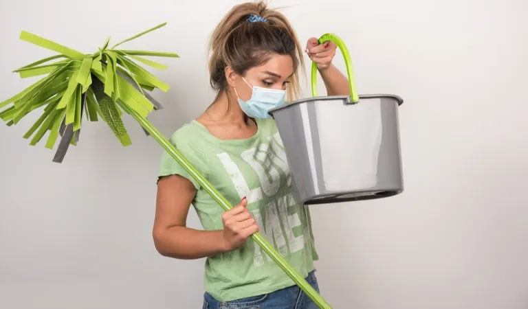 Lady in green top and mask holding a green stick and a basket in her hand.