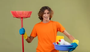 Young man wearing rubber gloves holding basin with cleaning tools and mop